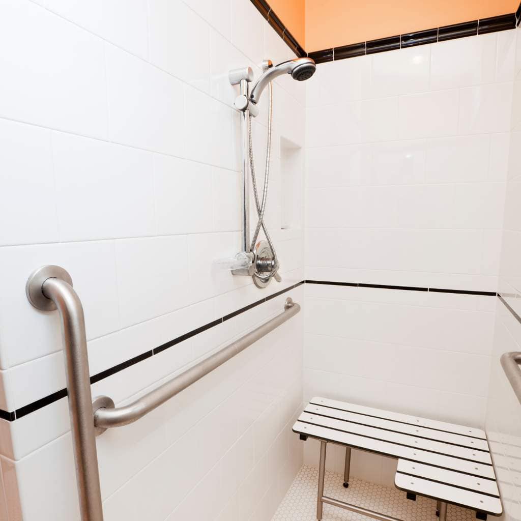 Accessible white-tiled roll-in shower with horizontal and vertical stainless steel grab bars, a handheld shower head, and a wall-mounted folding shower seat designed for ADA compliance.