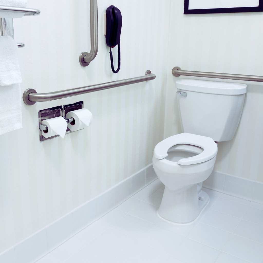 Elderly hand gripping a curved, stainless steel grab bar mounted on a light tile wall. The image emphasizes the secure grip and the required mounting height for safe user transfer near a toilet or shower.