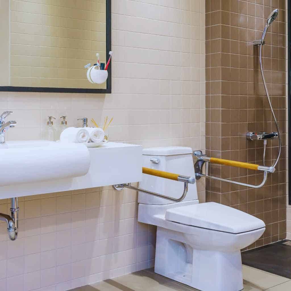 Accessible California bathroom with a wall-mounted sink, insulated piping, folding grab bars at the toilet, and open floor space illustrating ADA and Title 24 fixture dimension requirements.